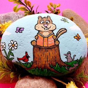 May include: A hand-painted rock depicting a squirrel sitting on a tree stump reading a book titled "Fairy Tales". The rock is painted with a blue sky, green grass, a red cardinal, and two mice.
