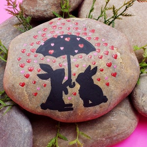 May include: A painted rock with two bunnies under an umbrella, surrounded by pink hearts. The bunnies are black and the umbrella is black with a white handle. The rock is brown and has a rough texture.