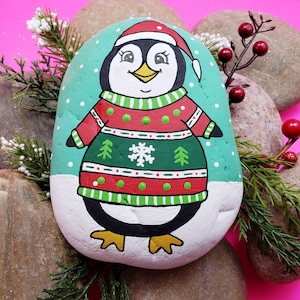 Könnte beinhalten: Handbemalter Stein mit einem Pinguin, der einen rot-grünen Weihnachtspullover mit Schneeflockenmuster trägt. Der Pinguin steht auf einem weißen Hintergrund mit blauem und weißem Schnee.