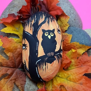 May include: A hand-painted rock with an orange background and a black silhouette of an owl perched on a tree branch. Two small black spiders are also painted on the rock.