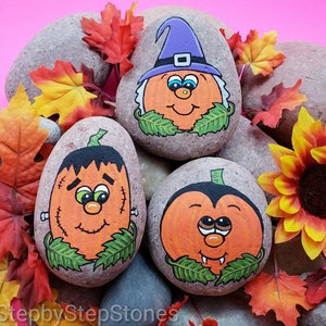 May include: Three hand-painted Halloween-themed rocks. One rock features a pumpkin wearing a witch hat, another a Frankenstein pumpkin, and the third a vampire pumpkin. The rocks are orange with green leaves and black details, surrounded by fall leaves.