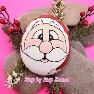 May include: A painted rock featuring a Santa Claus face. The rock is surrounded by small branches with white berries and red berries. The background is pink. The text "Step by Step Stones" is at the bottom of the image.