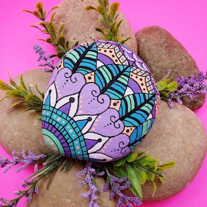 May include: A painted rock with a mandala design in shades of purple, blue, green, and orange. The rock is surrounded by small purple flowers and green leaves.