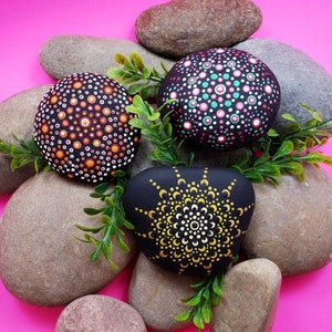 May include: Three painted rocks with mandala designs. The rocks are painted in black, white, orange, yellow, pink, and teal. The rocks are arranged on a pink surface with green foliage.