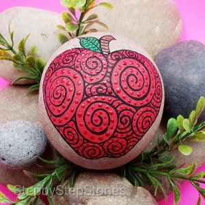 May include: A hand-painted red apple rock art with a swirl design and red glitter accents. The rock is surrounded by green foliage and other rocks.