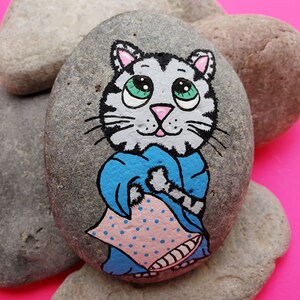 May include: Hand-painted rock art featuring a grey tabby cat with big green eyes, wearing a blue robe and holding a pink pillow with white polka dots.