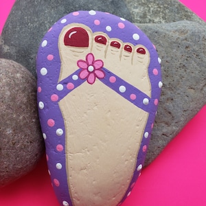 May include: A painted stone in the shape of a foot, featuring a purple sandal with white and pink polka dots. The toenails are painted red, and a pink flower decorates the sandal strap. The stone rests on a bright pink background.