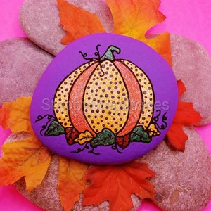 May include: A hand-painted purple rock featuring a yellow and orange pumpkin with black polka dots. The pumpkin is surrounded by green leaves and red and orange autumn leaves.