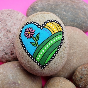 May include: A heart-shaped rock painted with a colorful landscape scene. The scene includes a blue sky, green hills, a yellow sun, and a pink flower. The heart is outlined in black with white dots.