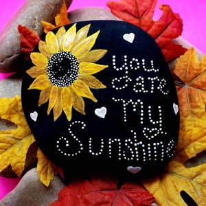 May include: A black painted rock with a yellow sunflower and white polka dots. The words "You are my sunshine" are written in white polka dots on the rock.