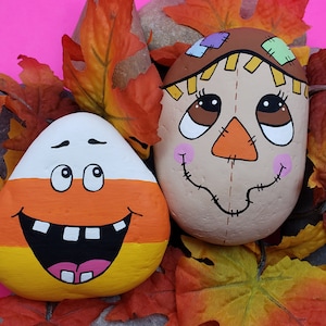 Puede incluir: Dos piedras pintadas, una con forma de caramelo de maíz con una cara sonriente y la otra con forma de espantapájaros con una sonrisa cosida. Ambas piedras están rodeadas de hojas de otoño.