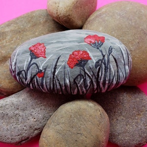 May include: A painted rock with a gray background and three red flowers with black stems and leaves. The rock is surrounded by other rocks.