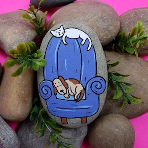 May include: Hand-painted rock art depicting a blue armchair with a white cat sitting on the back and a brown dog sleeping in the seat.