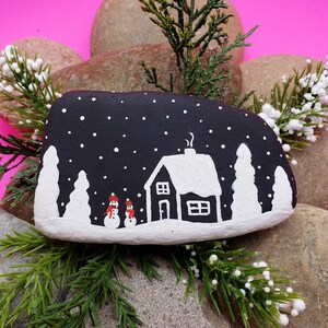 May include: A hand-painted, black stone featuring a winter scene. The design includes a white house with a snow-covered roof, two snowmen, and falling snowflakes. White trees are painted on either side of the house. The stone is surrounded by greenery.