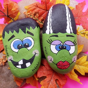 May include: Two painted rocks depicting cartoon monster faces. The rock on the left is green with a wide, toothy grin and black hair. The rock on the right is green with a purple eyelid, red lips, and white hair.