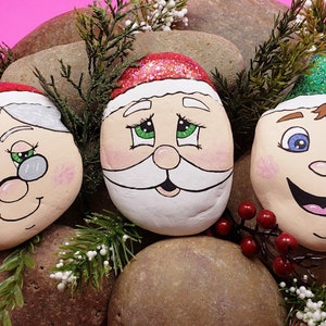 May include: Three painted rocks depicting Santa Claus, Mrs. Claus, and an elf. The rocks are decorated with glitter and have painted faces with festive expressions.