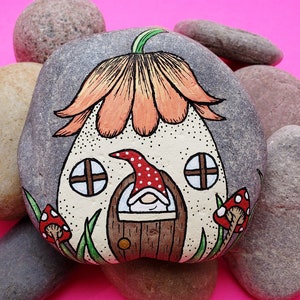 May include: Hand-painted rock art depicting a gnome's house with a flower-shaped roof, a brown door, and two windows. The gnome is peeking out of the door, wearing a red hat with white polka dots.