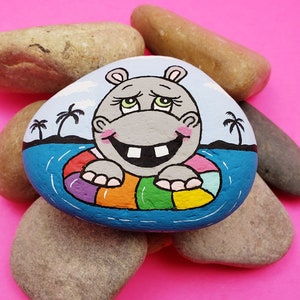 May include: Hand-painted rock art of a smiling gray hippopotamus wearing a rainbow inflatable ring in a blue pool. The background is a light blue sky with white clouds and palm trees.