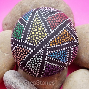May include: A black painted rock with a geometric design of colorful dots. The rock is surrounded by other rocks.