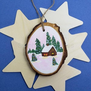 Op de afbeelding: Een handgeschilderd houten ornament met een klein huisje met een besneeuwd dak en ramen, omringd door dennenbomen in een winters landschap. De achtergrond is zacht roze.