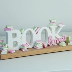 May include: A white decorative sign featuring the word BOOK in block letters and the word lover in cursive script, mounted on a rectangular wooden base. The sign is decorated with numerous miniature pastel pink and light green stacked books.