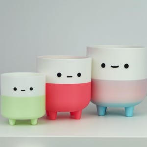 May include: Three ceramic plant pots with cartoon faces. The pots are white at the top, transitioning to green, red, and pink at the bottom. Each pot has three small legs and a simple black face design.