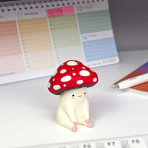 Può includere: Una piccola statuetta a forma di fungo dipinta a mano con un cappello rosso brillante a pois bianchi e un gambo color crema. Il fungo presenta un volto semplice disegnato a mano con due punti neri per gli occhi, una piccola bocca e guance rosa.