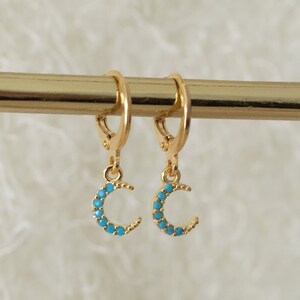 May include: Gold-coloured hoop earrings featuring crescent moon charms. Each moon is embellished with small, turquoise-coloured stones. The earrings are suspended from a gold bar, set against a soft, white backdrop.