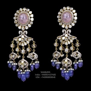 May include: A pair of gold earrings featuring pink and blue gemstones. The earrings have a round pink stone surrounded by smaller white stones, followed by a dangling chain of smaller white stones and blue stones.