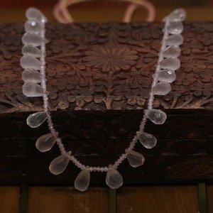 May include: A strand of pale pink rose quartz beads in a teardrop shape, displayed on a wooden box with intricate carvings.