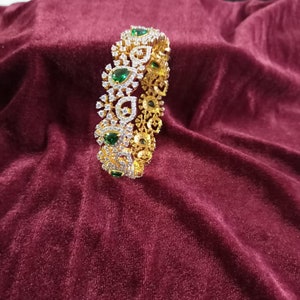 May include: A gold bangle bracelet with a decorative design featuring white stones and green gemstones. The bracelet is on a maroon velvet surface.