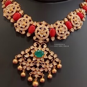 Pode incluir: Um colar de ouro com um pingente que apresenta uma gema verde cercada por pedras brancas. O colar é adornado com contas de coral vermelho e pedras brancas.