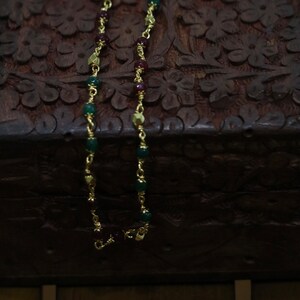 May include: A gold chain necklace with green and red beads. The necklace is on a dark brown wooden surface.