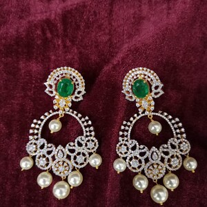 May include: A pair of gold earrings with a circular design featuring white stones and a green stone in the center. The earrings are accented with white pearls.