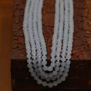 May include: A light blue beaded necklace made of round, faceted beads. The necklace is layered and is displayed on a wooden box.