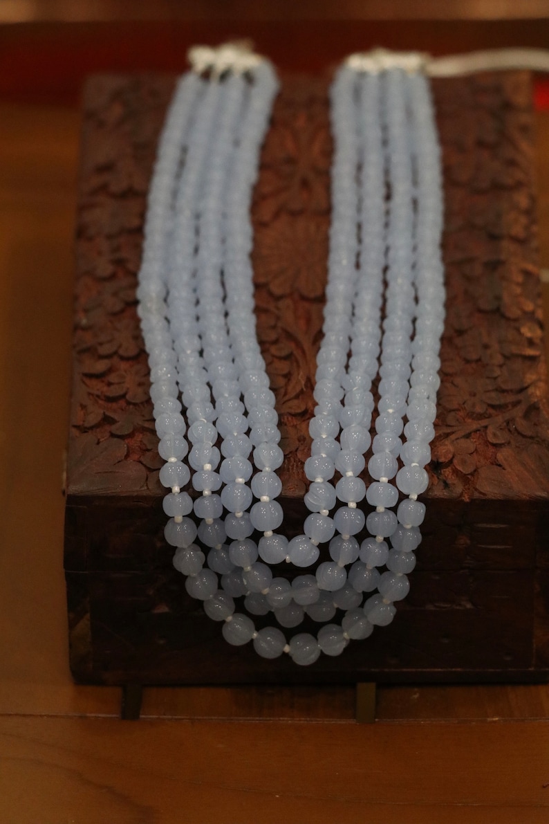 May include: A light blue beaded necklace made of round, faceted beads. The necklace is layered and is displayed on a wooden box.