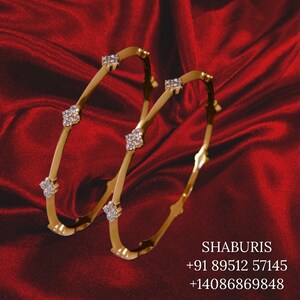 May include: Two gold bangles with diamond accents. The bangles are on a red fabric background.
