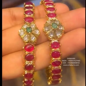 Pode incluir: Dois braceletes de ouro com pedras preciosas vermelhas e um design de flor branca com detalhes verdes. Os braceletes estão em uma mão.