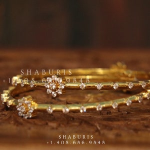 May include: Two gold bangles with a row of small clear stones around the edge. The bangles are stacked on top of each other and have a larger cluster of stones in the centre. The text "SHABURIS +1 408 686 9848" is visible in the image.