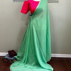 Könnte beinhalten: Ein hellgrüner, durchsichtiger Sari mit goldener Einfassung. Der Sari ist über eine Schaufensterpuppe drapiert, die ein leuchtend pinkfarbenes Oberteil mit weißen Punkten trägt.