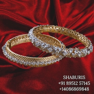 May include: Two gold bangles with diamond detailing are displayed on a red satin backdrop. The bangles showcase detailed patterns and are embellished with multiple small diamonds, producing a shimmering appearance. The text "SHABURIS" is visible.
