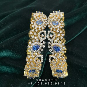 May include: Two gold bangles with intricate designs featuring blue gemstones and white diamonds. The bangles are set against a green velvet background.