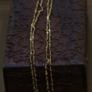 May include: A gold chain necklace with brown beads. The necklace is lying on a dark brown wooden box with intricate carvings.
