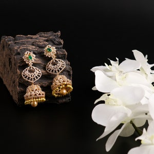 May include: A pair of gold earrings with a teardrop design, featuring a green stone at the top and a pearl-like drop at the bottom. The earrings are set against a dark background with a piece of wood and white flowers.
