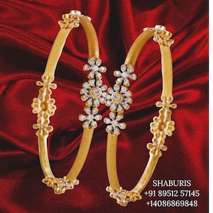 May include: Two gold bangles with floral and diamond detailing. The bangles have a matte finish and are set against a red background. The floral designs are clustered in the centre and at the ends of the bangles.