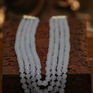 May include: A multi-strand necklace made of light blue beads. The beads are round and have a slightly textured surface. The necklace is displayed on a wooden box.