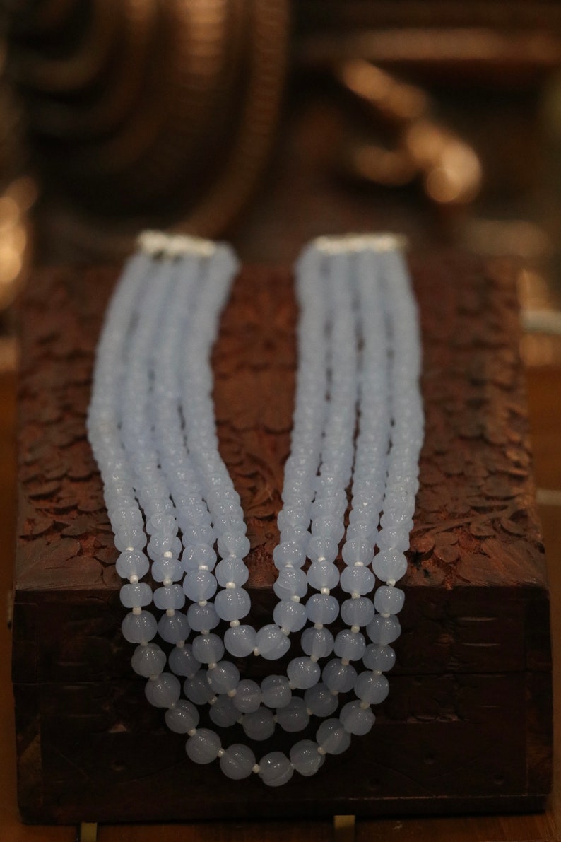 May include: A multi-strand necklace made of light blue beads. The beads are round and have a slightly textured surface. The necklace is displayed on a wooden box.