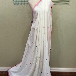 Könnte beinhalten: Weißer, durchsichtiger Sari aus Stoff mit rosa Saum und kleinen runden Spiegelverzierungen.