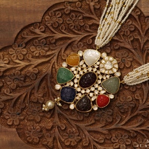 Puede incluir: Un collar de oro con un gran colgante redondo que presenta una variedad de piedras preciosas de colores, incluyendo verde, azul, rojo y morado. El colgante está rodeado de piedras preciosas más pequeñas y transparentes. El collar está hecho de perlas blancas y cuentas doradas.