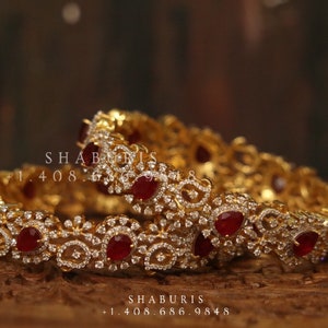 May include: Two gold bangles featuring intricate designs and red gemstones. The bangles are set with small, clear stones that create a sparkling effect.